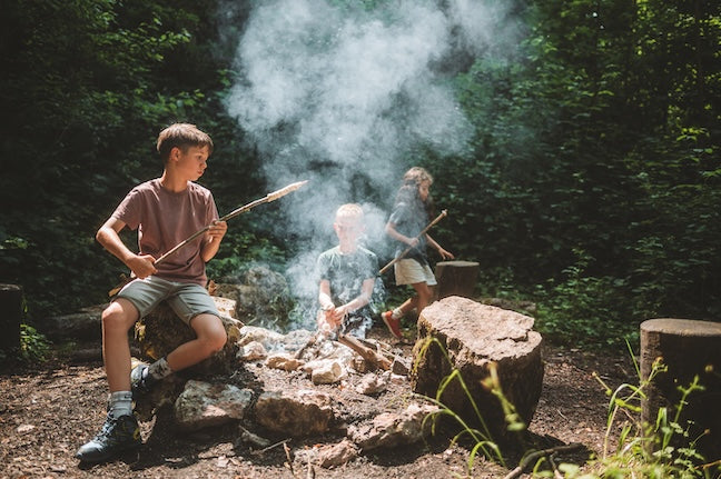 Drei Kinder sind im Wald am Schlangenbrot grillieren über einem Feuer.