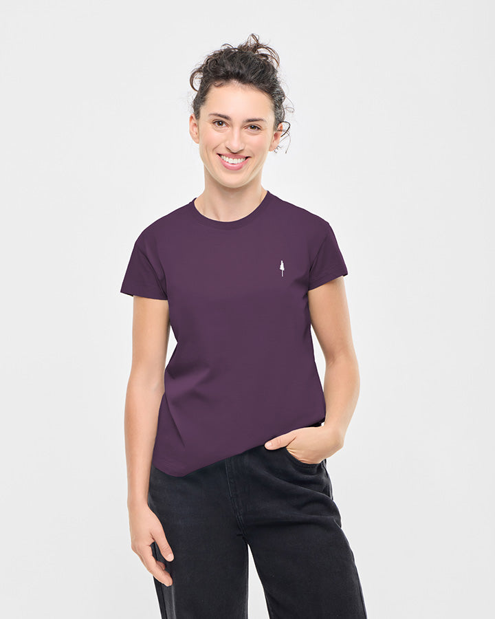 A person with curly hair in a bun is smiling and wearing a plain deep purple TreeShirt Women T-shirt with a small white rocket icon on the left chest, paired with black pants, standing against a light gray background.