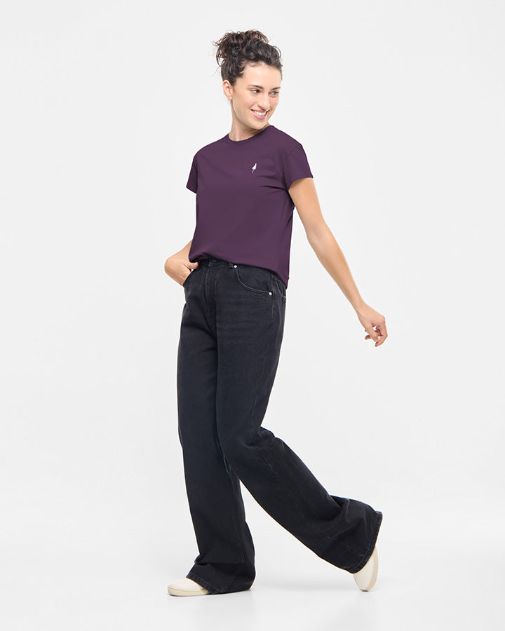 A person with curly hair wears a TreeShirt Women Deep Purple, loose black jeans, and white shoes, standing against a plain white background and smiling while looking to the side.