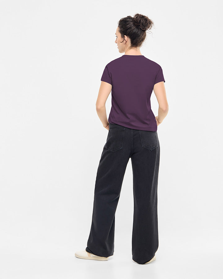 A woman stands facing away, wearing a TreeShirt Women Deep Purple with short sleeves, loose black jeans, and beige shoes. Her hair is tied up against a plain white background.