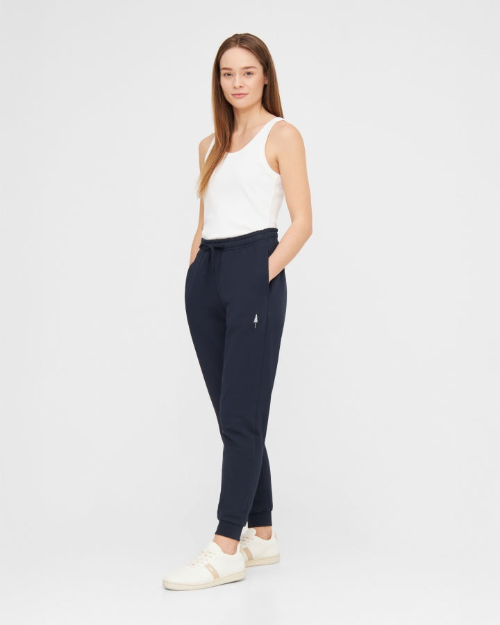 NIKIN Ensemble de survêtement pour femme : débardeur blanc et pantalon de jogging bleu marine, les mains dans les poches, sur fond blanc.