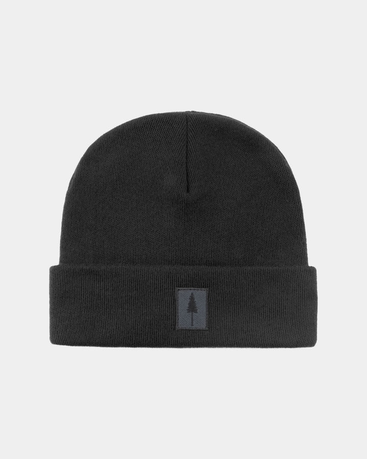 The Nature Nomad Treeanie is a plain black knit beanie with a folded brim and a small square patch showing a simple tree design on the front. This eco-friendly accessory is displayed against a light gray background.