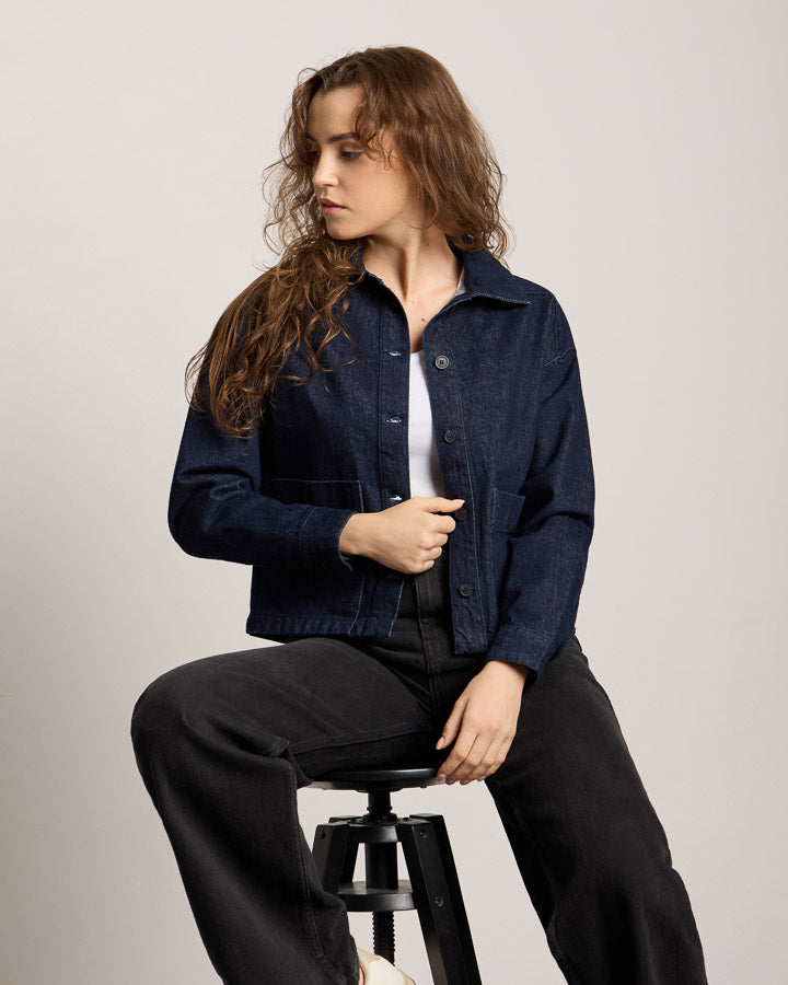 NIKIN TreeJacket Denim ReCotton Damen-Jeansjacke, dunkles Indigo, auf einem Hocker sitzend, neutraler Hintergrund.