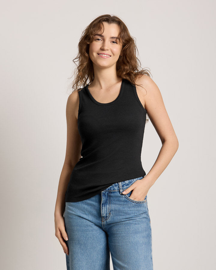NIKIN  TreeTop Women Damen-Shirt, schwarzes Oberteil, blaue Jeans, lässig mit einer Hand in der Tasche, heller Hintergrund.