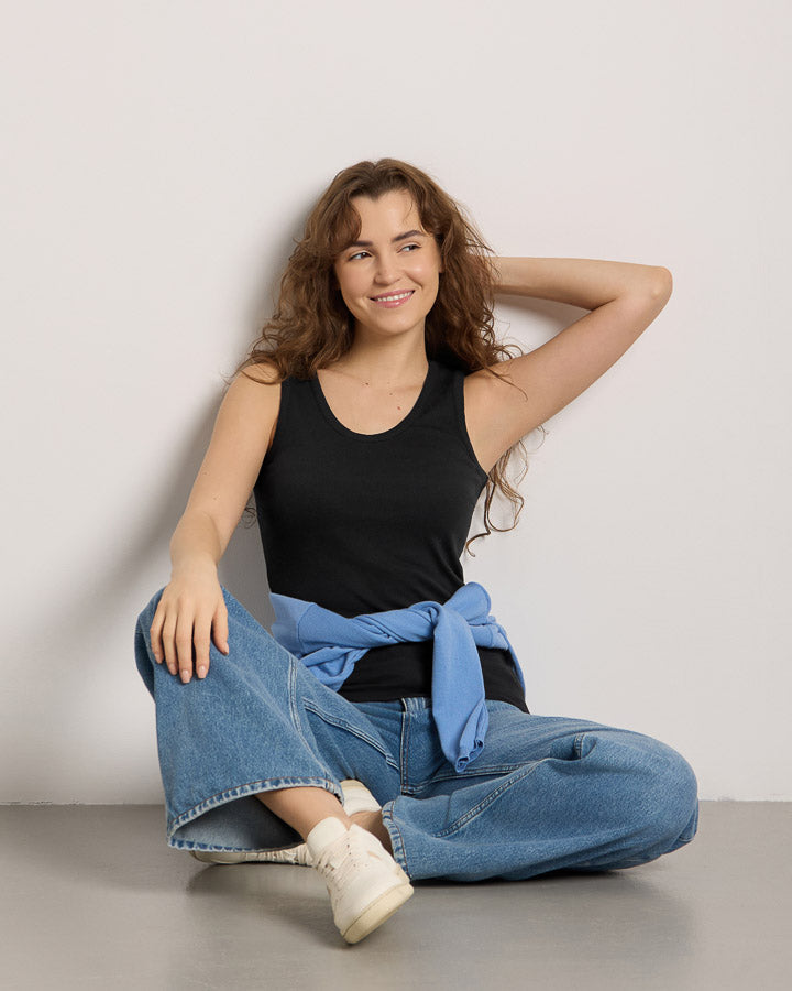 NIKIN TreeTop Women Damen Top schwarz, kombiniert mit Jeans, Model sitzt am Boden und lächelt.