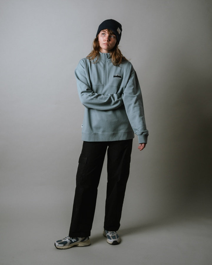 NIKIN  TreeSweater Quarter Zip Muntogna Pullover, Elemental Blue, Herrenmode, schwarze Hose, Sneakers, Beanie, neutraler Hintergrund.