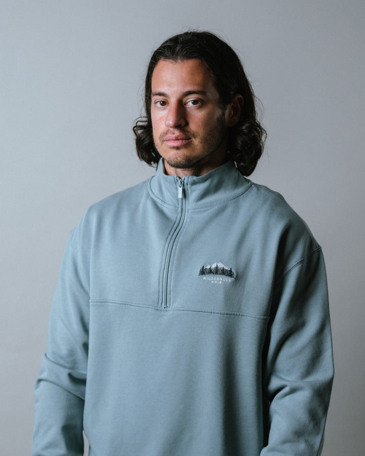 NIKIN  TreeSweater Quarter Zip Muntogna Herrenpullover, Elemental Blue, langes dunkles Haar, grauer Hintergrund.