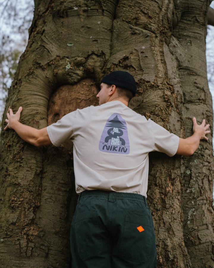 NIKIN TreeShirt Jubiläums-T-Shirt „Relaxed Pumice“, Treehugger-Aufdruck auf dem Rücken, Person mit Mütze umarmt einen Baum.