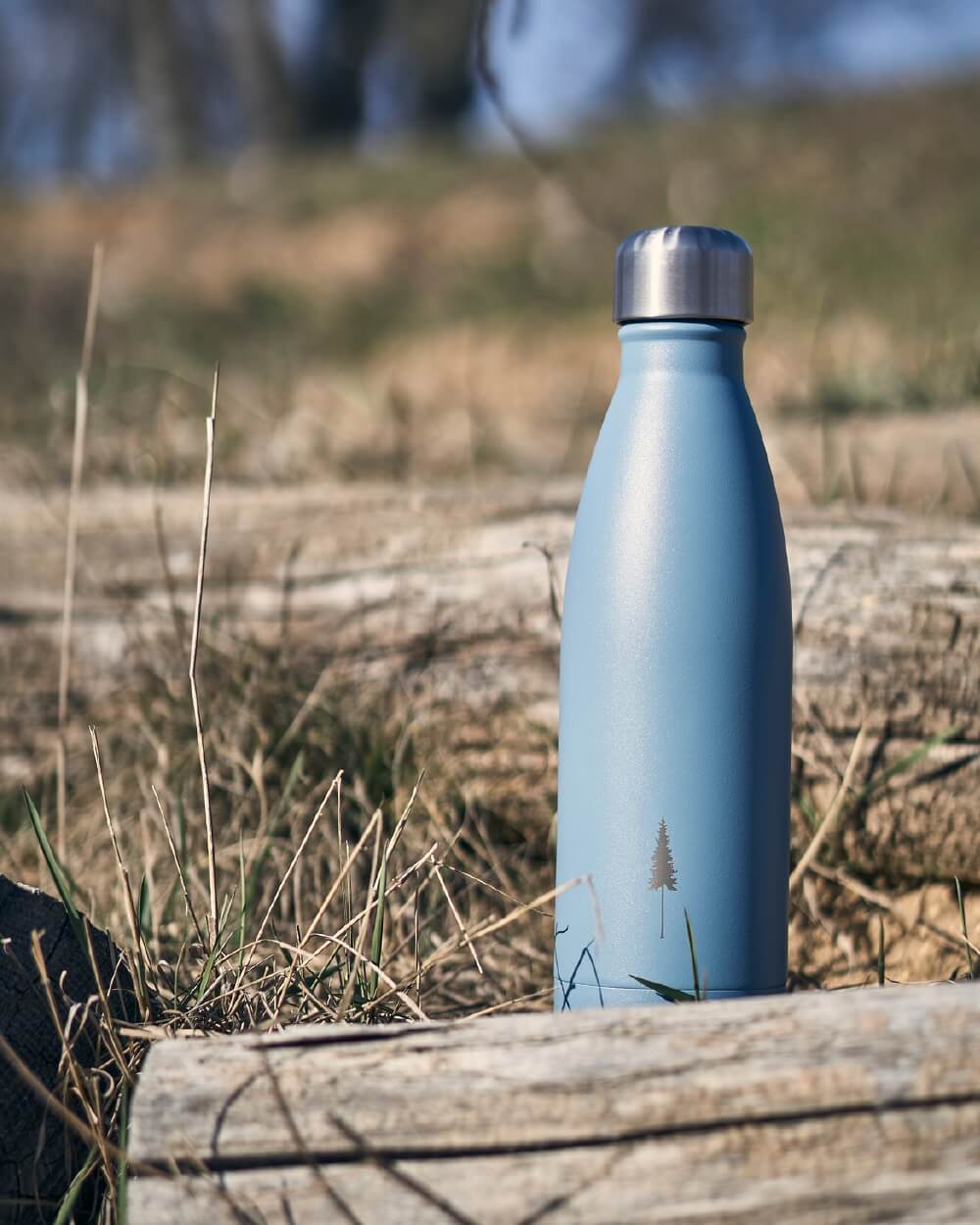 NIKIN TreeBottle hellblaue Trinkflasche mit Baum-Symbol, im Freien auf trockenem Gras und Holz platziert.