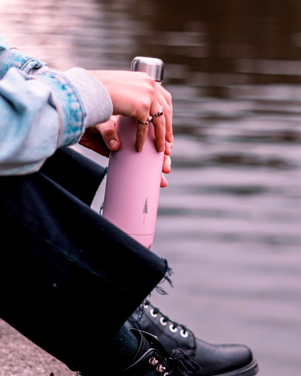 NIKIN Gourde TreeBottle Astrodust rose avec motif d'arbre, représentant une personne au bord de l'eau portant des bottes noires et un jean.